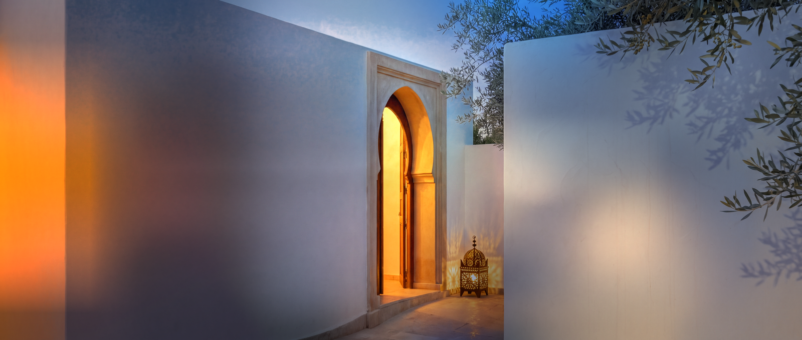 Caméra IP bullet Longse 12 MP blanche fixée sur le mur blanc d'une villa marocaine à l'heure bleue, ses deux LED allumées éclairant le plâtre, près d'une porte en arc sculpté ouverte sur une lumière ambrée et d'une lanterne en laiton